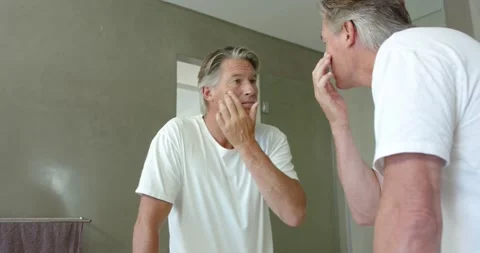 50s man standing at bathroom vanity in white T-shirt, noticing mirror, checking Stock Footage 335574378