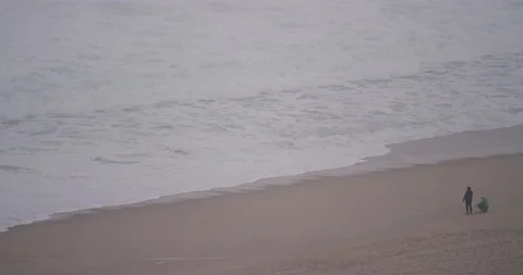 5K distant view of two surfers resting on sandy beach watching wavy ocean Stock Footage 157718064
