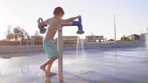 6 year old boy at splash pad plays with two rotating spray nozzles in summer Stock Footage 233939543