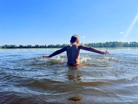 A 6-year-old stands in the river, waist-deep, and joyfully splashes around. Stock Photos
