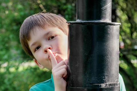 8-year-old boy with dark hair stands behind a metal lamppost and shows gestur Stock Photos