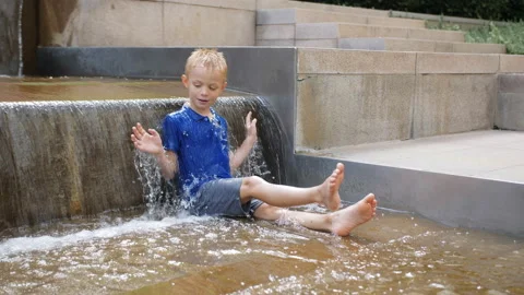 An 8-year-old boy is sitting in a city fountain under flowing streams of water. Stock Footage 205597366