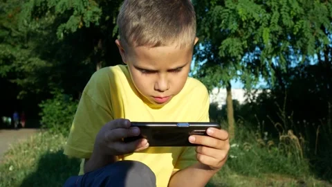 An 8-year-old boy squatted down and plays a game on his smartphone. The guy is Stock Footage 309176976