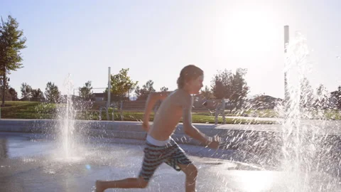 9 year old boy jumps through splash pad water jet in slow motion Stock Footage 233938424