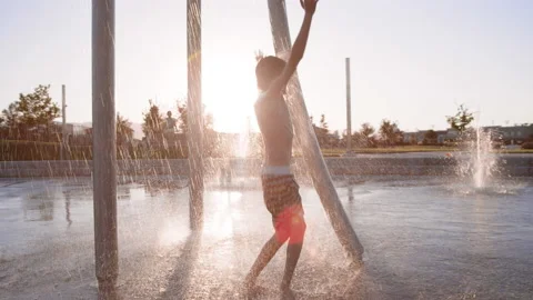 9 year old boy jumps and dances under raining water at splash pad, slow motion Stock Footage 233947258