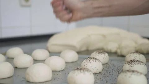 A4K. Baker sprinkling a tray of bread rolls with seeds with dough behind Stock Footage 251363686