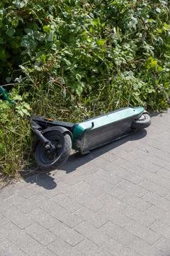 Abandoned e-scooter lying in grass by sidewalk Stock Photos