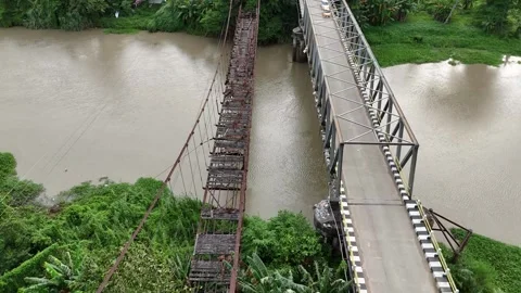 An abandoned old suspension bridge over a murky river surrounded by green trees. Stock Footage 324548149