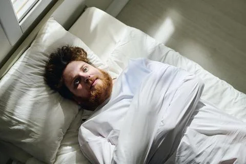 Above angle of young bearded man in straitjacket lying on bed in ward Stock Photos
