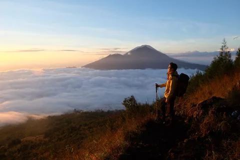 Above Cloud Nine, Mount Batur's Peak, Asian Man Trekker under Azure Sky and.. Stock Photos