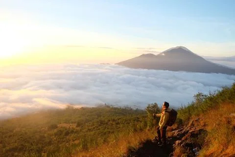 Above Cloud Nine, Mount Batur's Peak, Asian Man Trekker under Azure Sky and.. Stock Photos