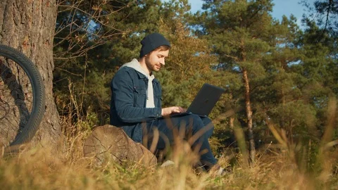 Active and mobile young man with laptop sitting in a clearing and networking in Stock Footage 99364861