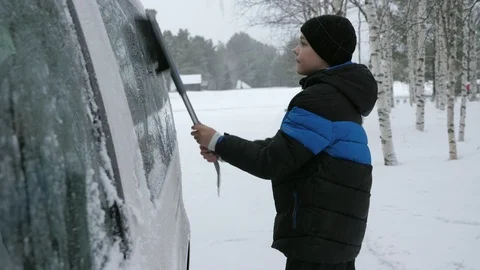 Active boy removing snow from a side window of a car in a snowy forest in Video stock 123088771