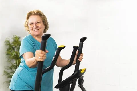 Active elderly woman smiles while exercising on a bicycle in the gym. Stock Photos