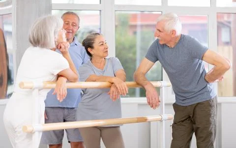 Active old attendees of ballet courses communicating cheerfully at ballet-barre 스톡 사진