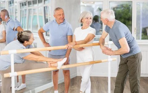 Active old attendees of ballet courses communicating cheerfully at ballet-barre 스톡 사진