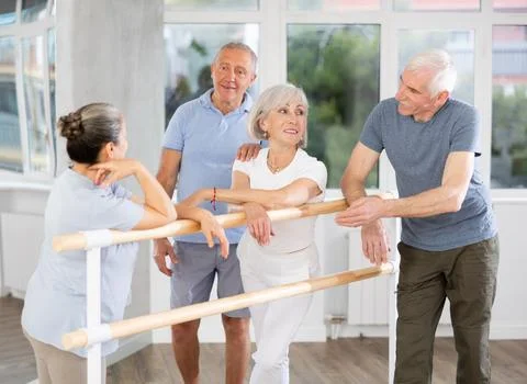 Active old attendees of ballet courses communicating cheerfully at ballet-barre 스톡 사진