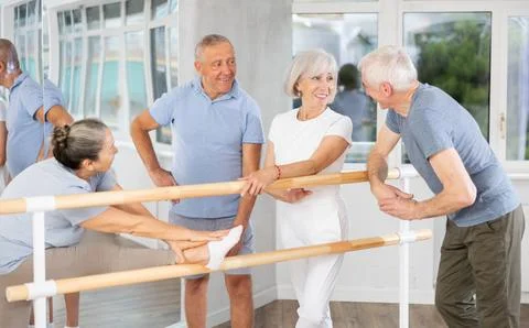 Active old attendees of ballet courses communicating cheerfully at ballet-barre 스톡 사진