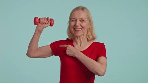 Active senior woman posing with dumbbells in hands. points fingers at muscles Stock Footage 261887853