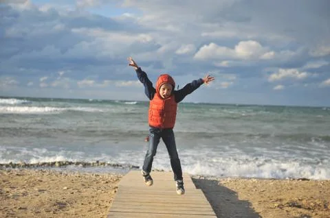 Active small boy in red and blue jacket and hood jumping near stormy sea in a Stock-Fotos