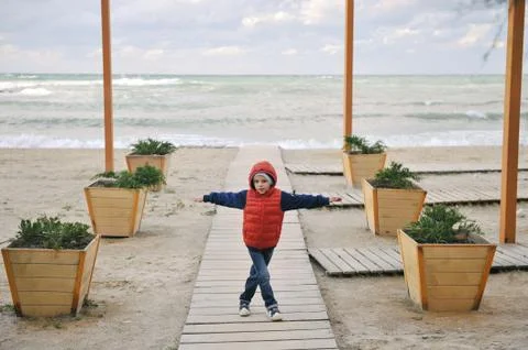 Active small caucasian boy in jacket with hood standing on empty sea beach in Foto stock