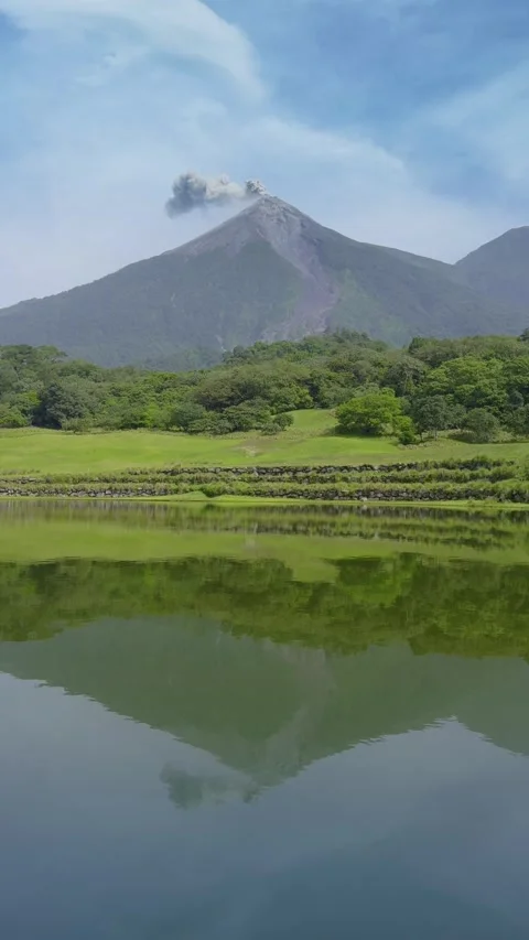 Active Volcano Emitting Smoke with Lake Reflection and Lush Green Landscape. Stock Footage 338360428