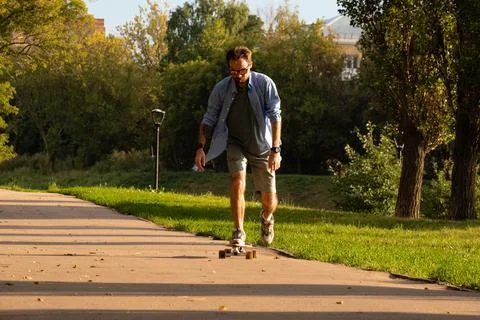 Active young man skating on longboard along empty asphalt roadway among trees Stock Photos