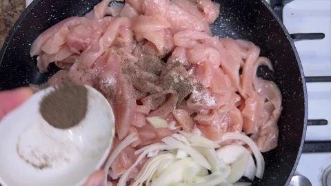 Adding black ground pepper to pieces of raw chicken and onions in a frying pan Stock Footage 301329099