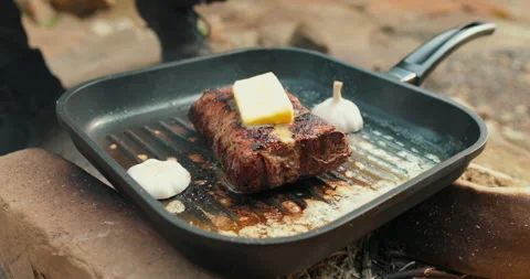 Adding butter to a steak on a hot pan in the process of grilling meat outdoors. Stock Footage 265151886
