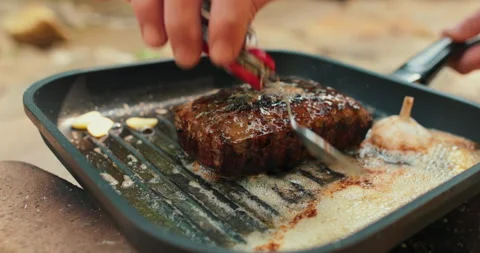 Adding butter to a steak on a hot pan in the process of grilling meat outdoors. Stock Footage 265152613