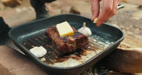 Adding butter to a steak on a hot pan in the process of grilling meat outdoors. Stock Footage 265153466