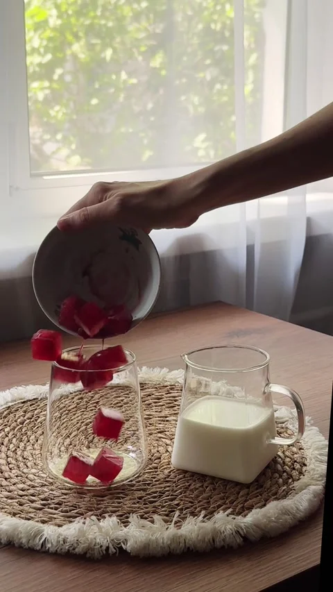 Adding cherry juice ice cubes to a clear glass next to a glass milk pitcher Video stock 341133960
