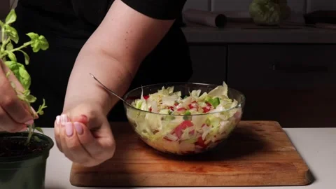 Adding fresh basil leaves to vegetable salad in glass bowl. Cooking process on Stock Footage 316472069