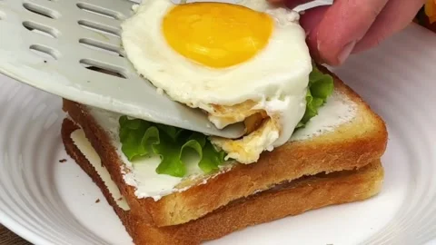 Adding a fried egg and fresh lettuce on a double layer toast sandwich Stock Footage 324943444