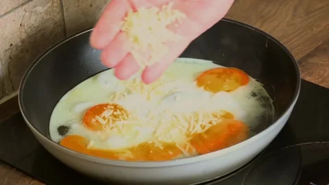 Adding grated cheese to frying eggs in hot pan during morning breakfast cooking Stock Footage 343258894