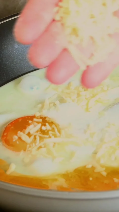 Adding shredded cheese to frying eggs in pan for breakfast recipe Stock Footage 343258839