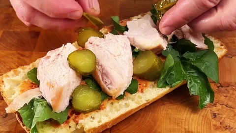 Adding sliced pickled cucumbers on chicken breast sandwich with arugula and sauc Stock Footage 325032703