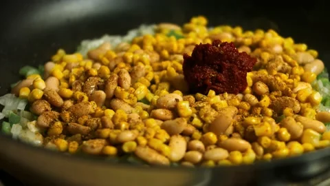 Adding tomato paste and water to beans, corn and chopped vegetables being fried Stock Footage 151290091