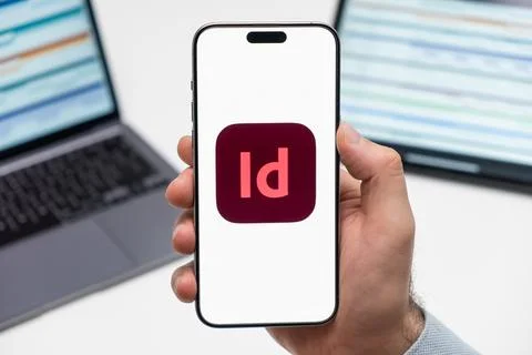 Adobe InDesign CC logo on the screen of smartphone in mans hand on the workplace Stock Photos