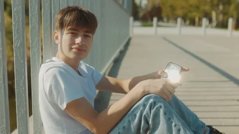 Adolescent boy sitting down outdoors, holding a smartphone in a sunny park Vídeo Stock 331250474