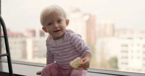 An adorable blonde baby joyfully eats a piece of bread while sitting on a high Vidéo 313638280