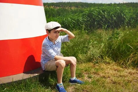 Adorable child boy sitting on the background of a bright lighthouse and looki Stock Photos