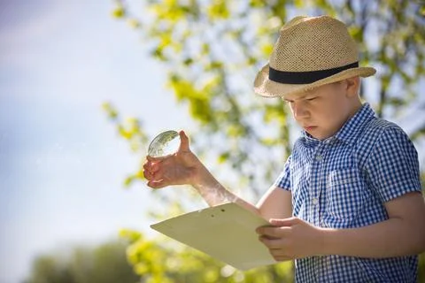 Adorable kid boy making fire on paper with a magnifying glass outdoors, on su Stock Photos