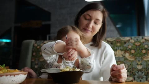 Adorable toddler learning to use fork with mother's help in restaurant Stock Footage 303777071