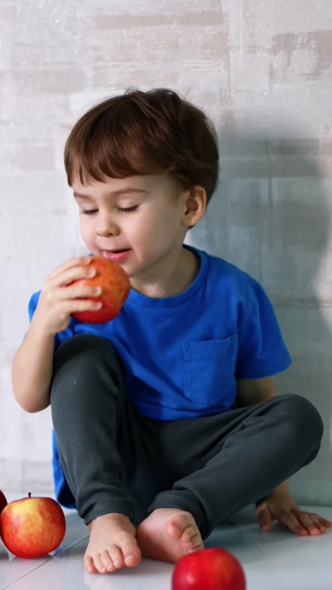 Adorable toddler sits surrounded by red apples. Cute baby boy picks one apple Stock Footage 333472589