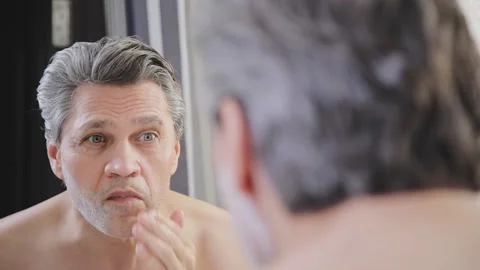 An adult gray-haired man applies shaving cream to his face in front of a mirror. Stock Footage