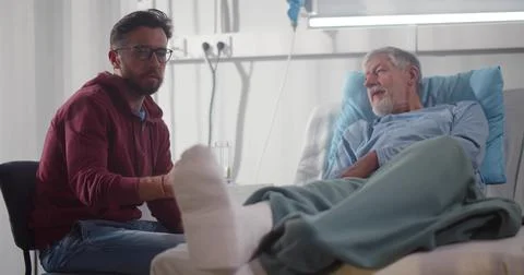 Adult man visiting aged father with broken leg in hospital ward Stock Photos