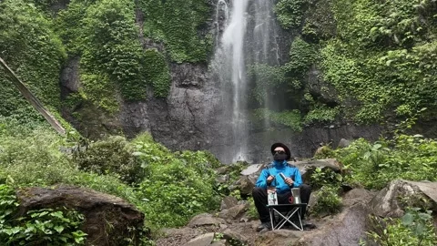 Adventurer Preparing Drink at Base of Lush Jungle Waterfall, Wide Shot Stock Footage 324850575