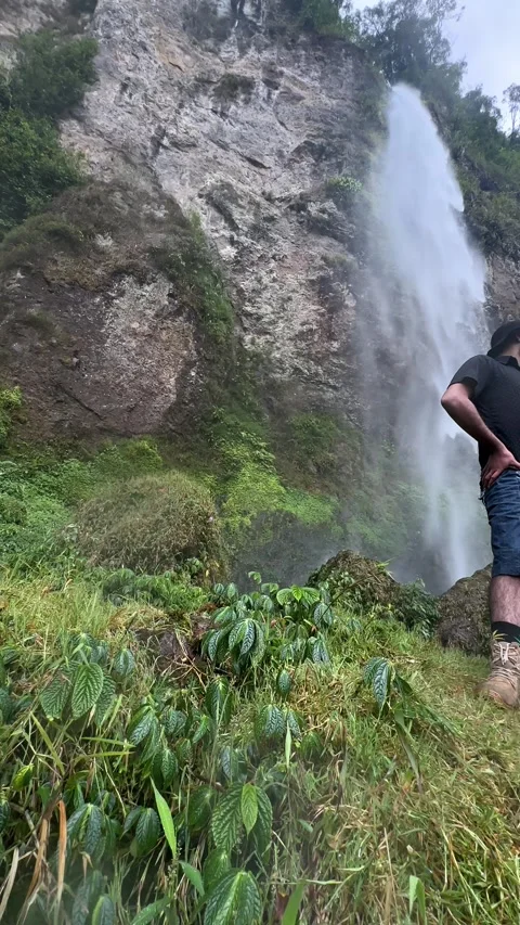 Adventurous Man Posing by Tall Waterfall, Rocky Cliff, Low Angle Vertical Shot Stock Footage 323912759