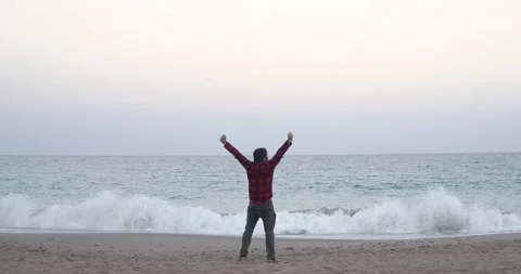 Adventurous man runs along the beach in casual clothes and raises his arms as Stock Footage 153597754